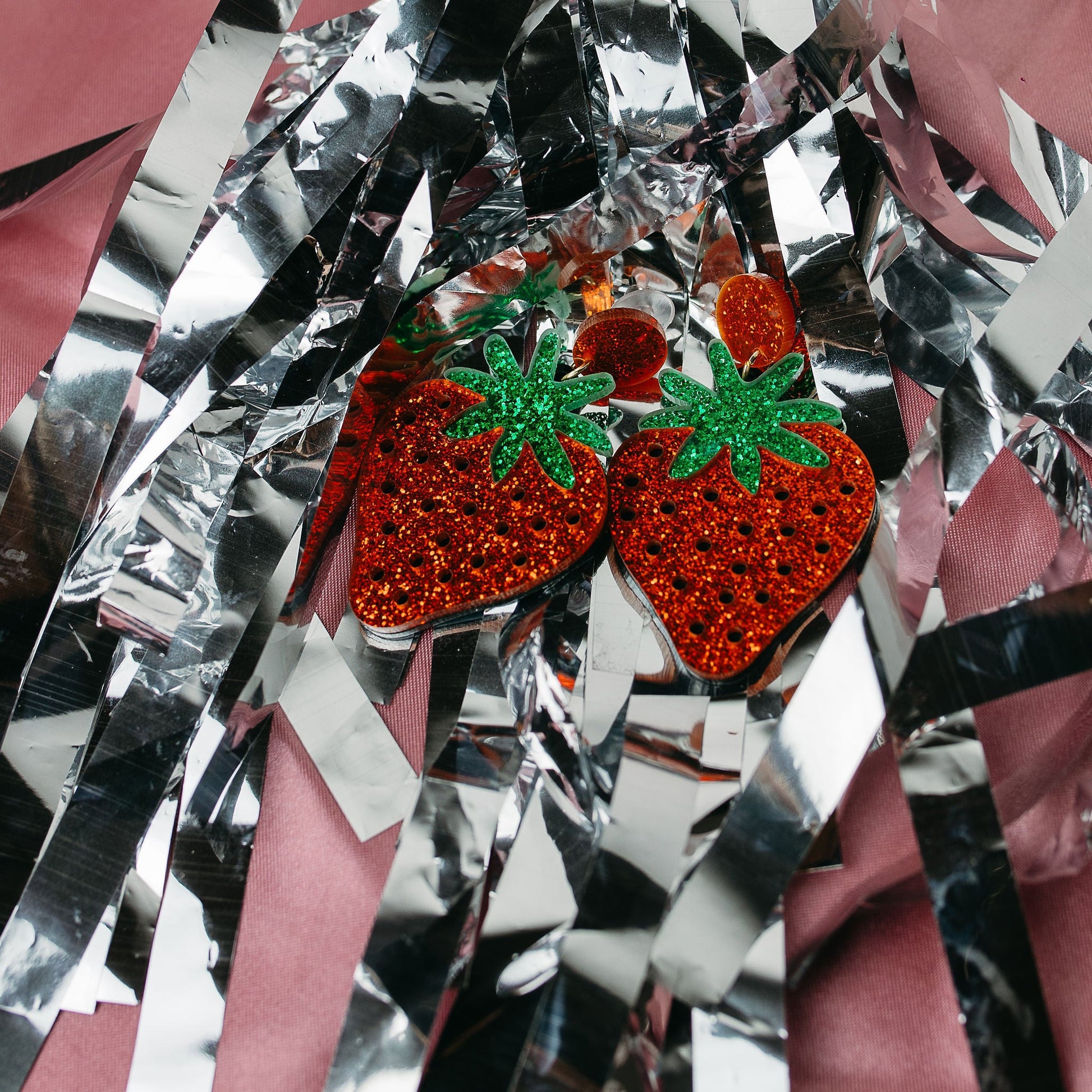 Strawberry-shaped earrings with red and green details on a reflective surface
